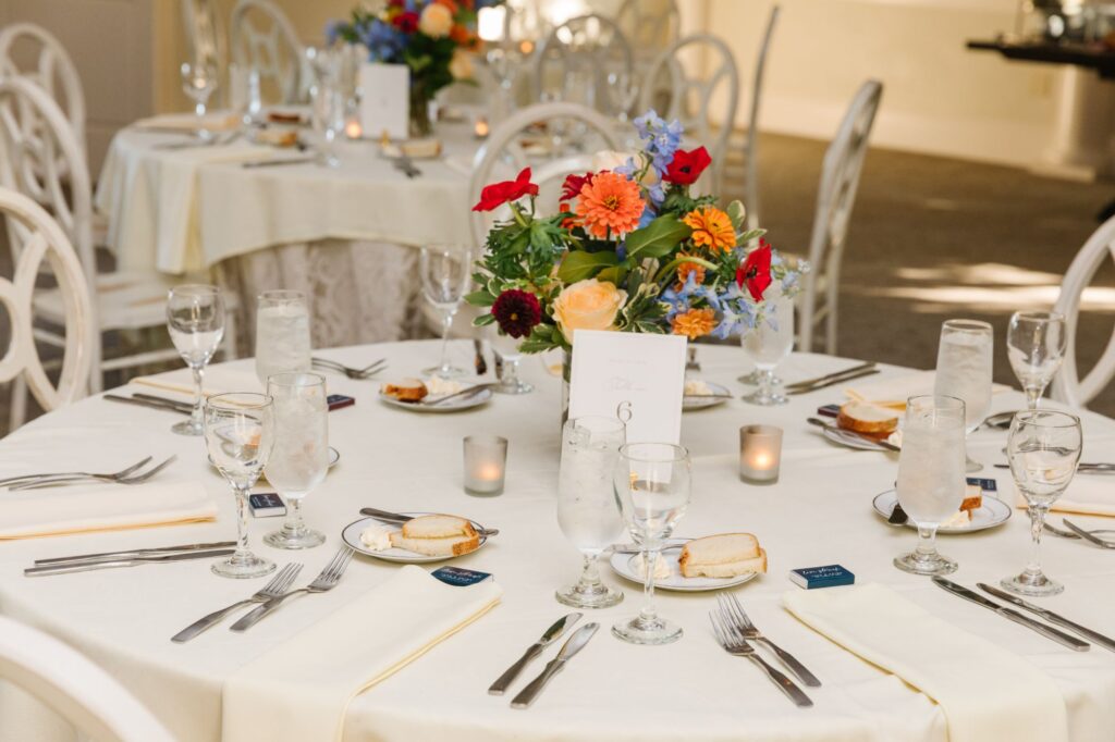 A round table set for a formal Chastain Horse Park rehearsal dinner, featuring a white tablecloth, floral centerpiece, glassware, plates with bread, silverware, and place cards. Other similarly dressed tables can be seen in the background.