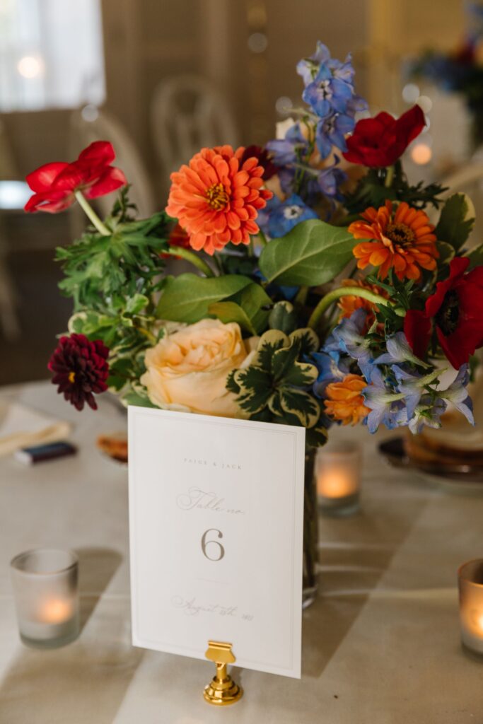 A colorful floral centerpiece with orange, red, blue, and cream flowers in a glass vase sits on a table at the Chastain Horse Park rehearsal dinner. In front, a card reads “Table 6,” surrounded by small votive candles for added charm.