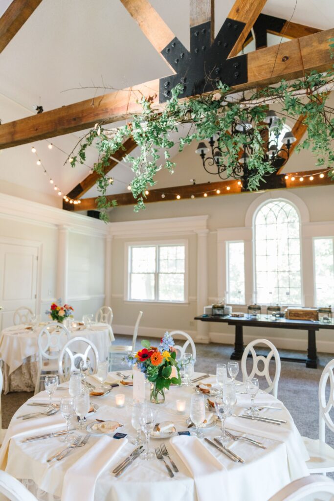 A bright, elegant event space at Chastain Horse Park with round tables set for dining, white chairs, and floral centerpieces. Wooden ceiling beams are adorned with greenery and string lights, while large windows fill your rehearsal dinner with natural light.