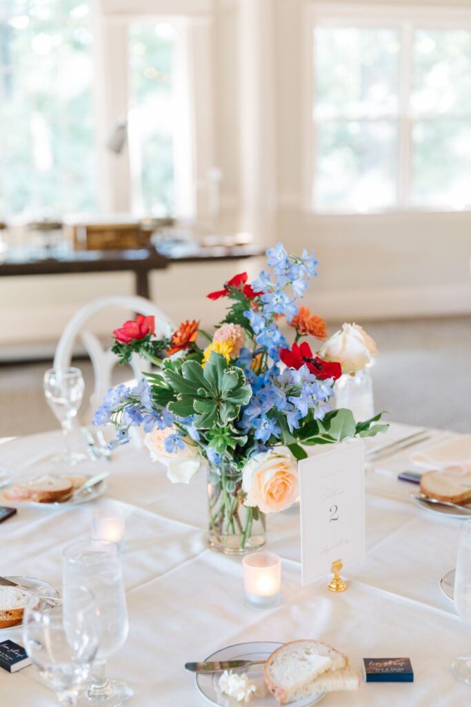 A bright floral centerpiece with blue, red, and peach flowers sits on a white-clothed table set with glassware, bread, candles, and a table number card at a sunlit Chastain Horse Park rehearsal dinner.