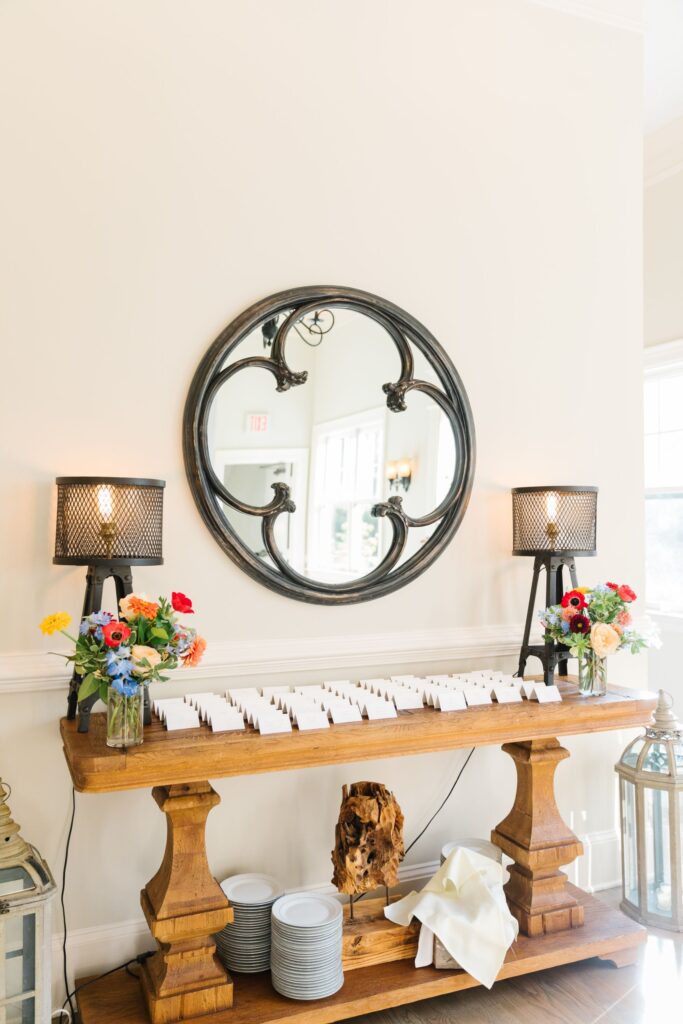 A wooden table with neatly arranged name cards, colorful flower vases, and two black lamps sits below a round mirror on a cream wall—perfect for a Chastain Horse Park rehearsal dinner. Plates and napkins are stored on the lower shelf.