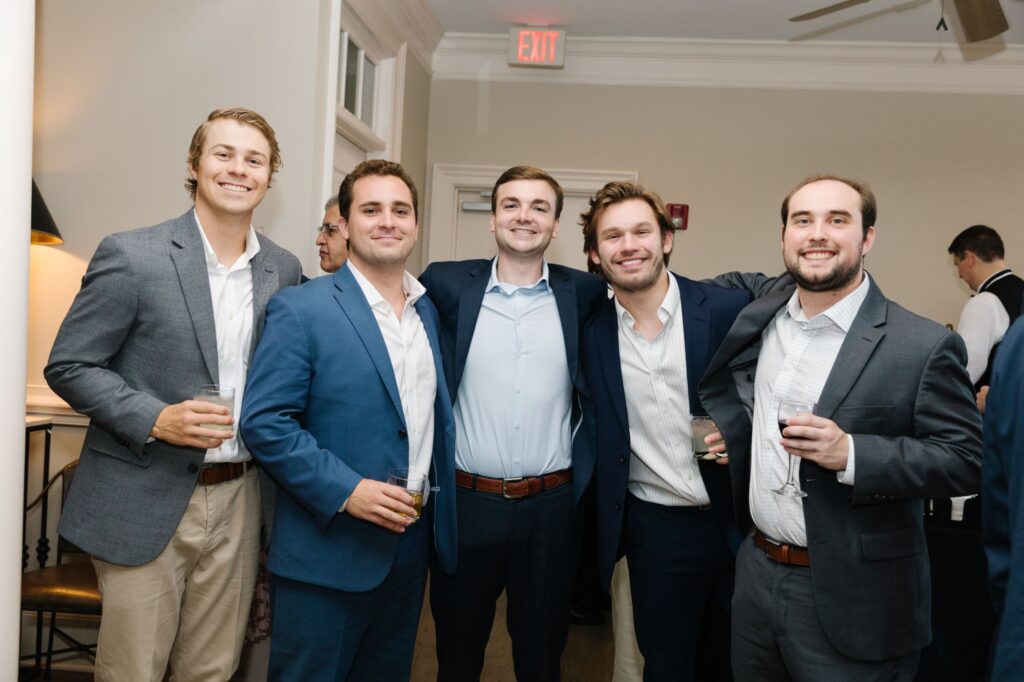 Five men in suits stand close together, smiling at the camera while holding drinks at a Chastain Horse Park rehearsal dinner. The background shows beige walls, a door, and an EXIT sign above them.