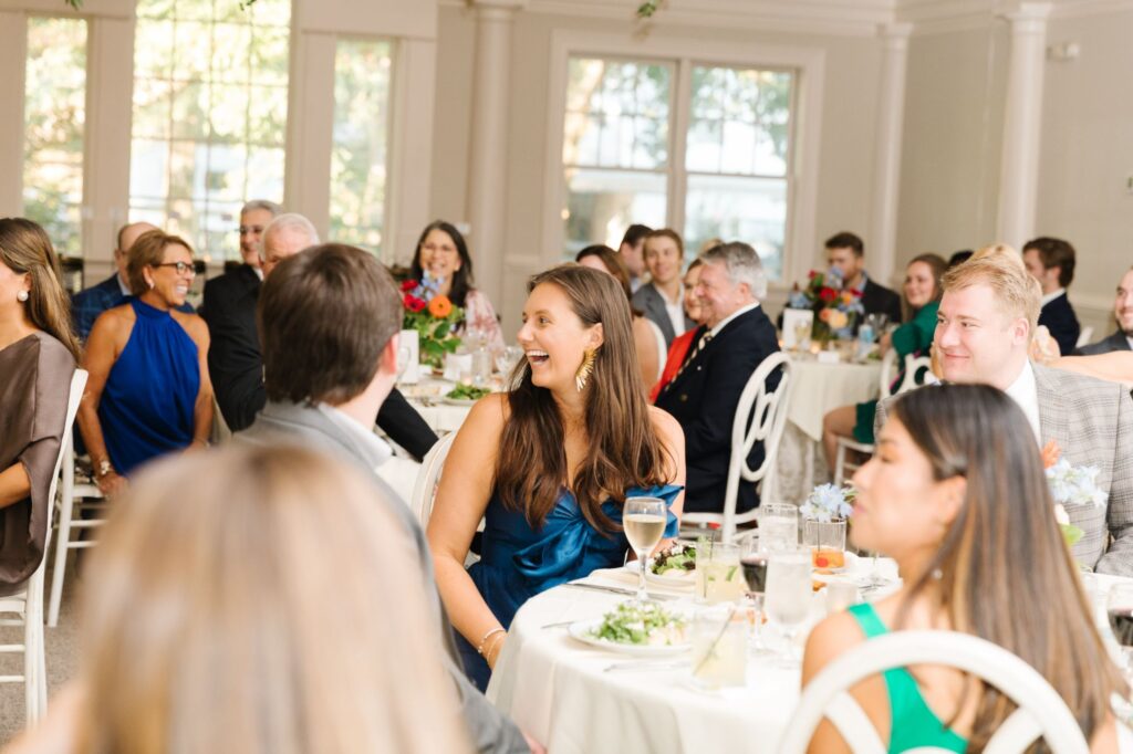 A group of people dressed in formal attire sit at round tables, smiling and talking over plates of food and drinks at a bright, elegant Chastain Horse Park rehearsal dinner event space.