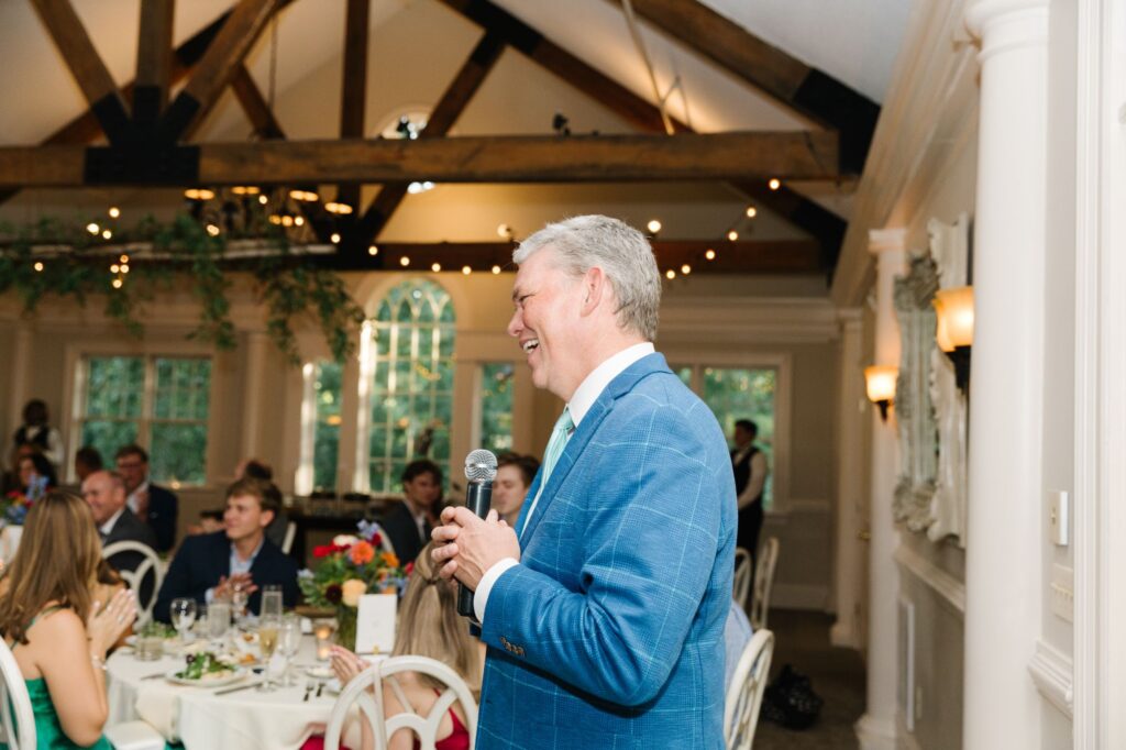 A man in a blue suit holds a microphone and speaks, smiling, at a Chastain Horse Park rehearsal dinner—a formal indoor event with seated guests and elegant decor including wooden beams and string lights.