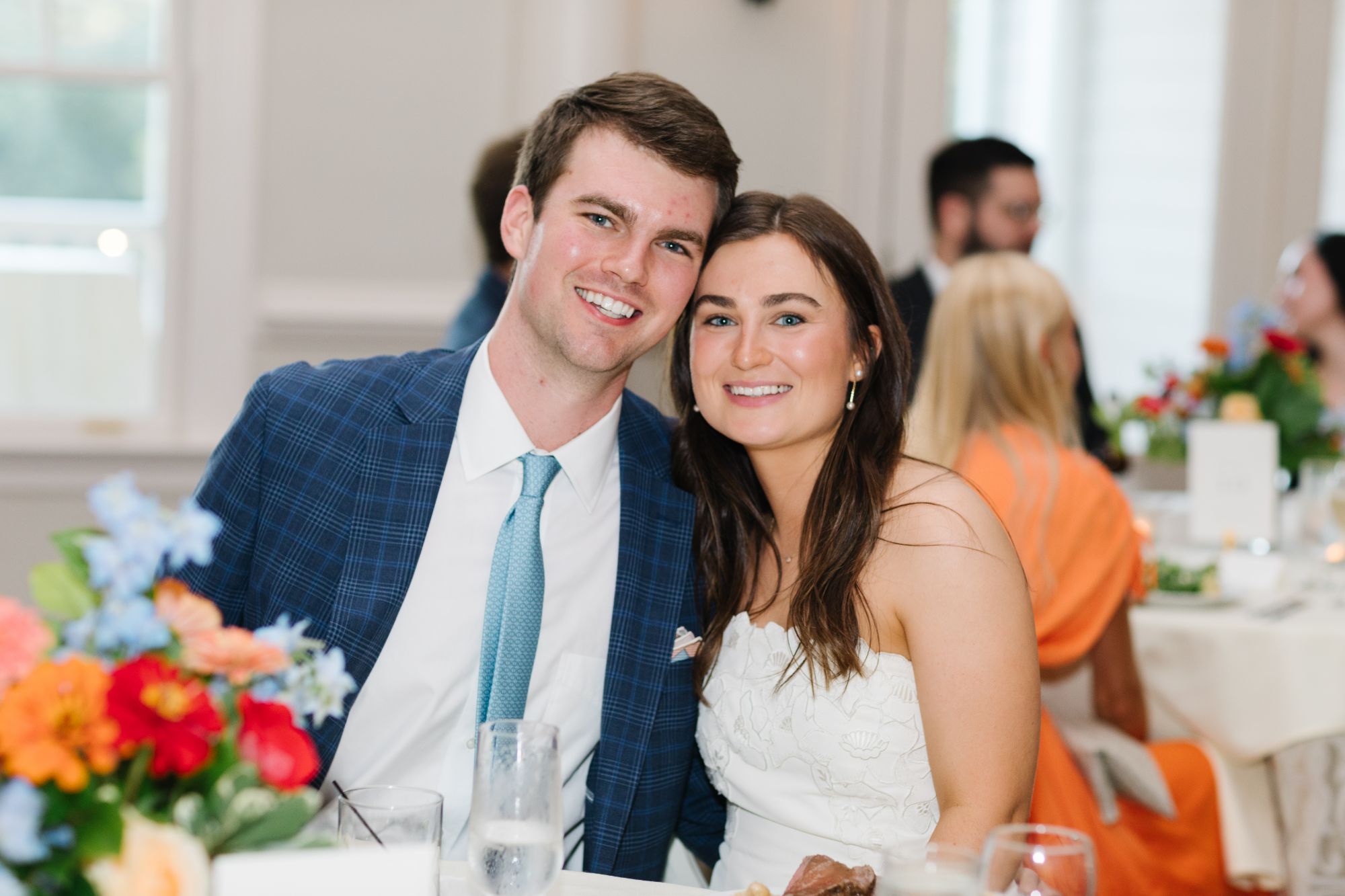 A smiling couple sits closely together at a decorated table during a Chastain Horse Park rehearsal dinner. The man wears a blue suit and tie; the woman wears a white dress. Colorful flowers and other guests are visible in the background.