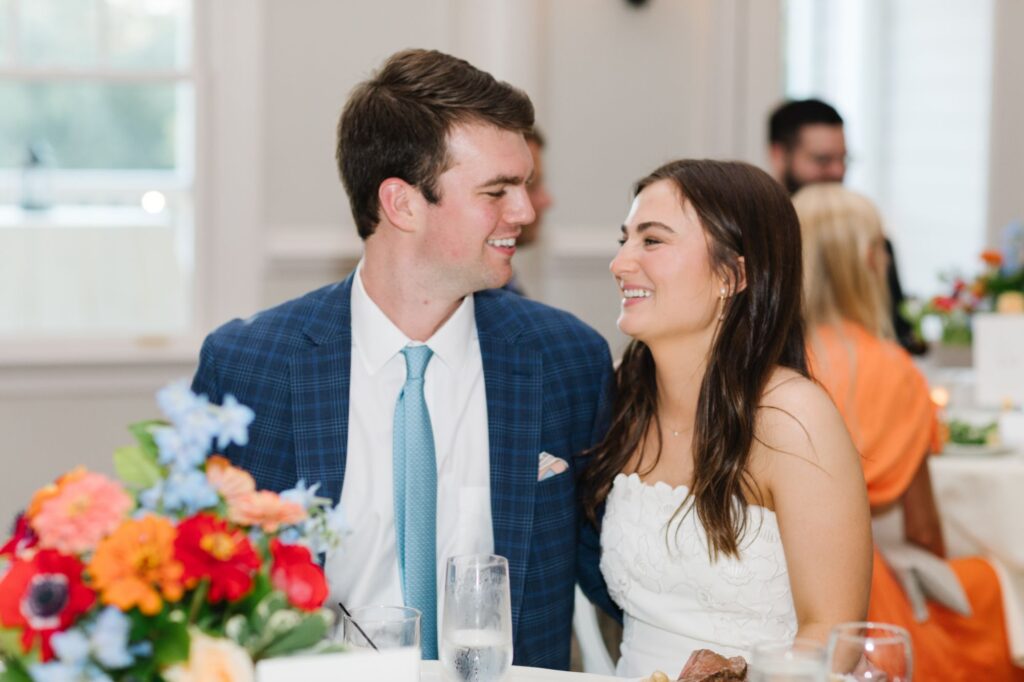 A smiling couple in formal attire sit closely together at a table decorated with colorful flowers, looking at each other lovingly during their Chastain Horse Park rehearsal dinner celebration.