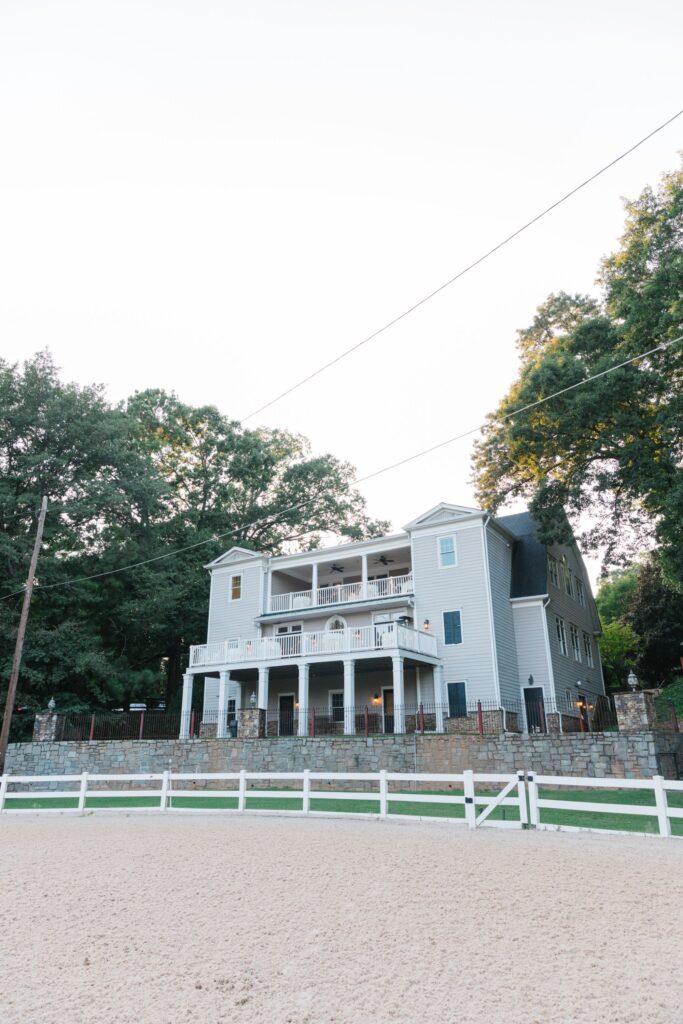 A large, light gray, three-story house with white trim and balconies is surrounded by trees and a stone wall, set behind a white fence and sandy yard—perfect for hosting a Chastain Horse Park rehearsal dinner under a clear sky.