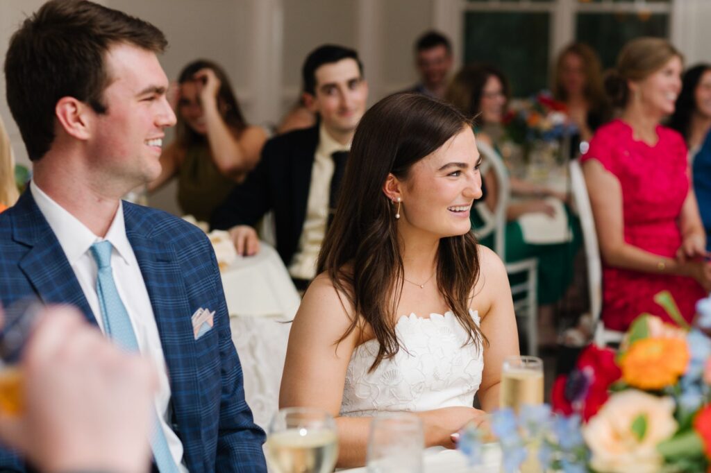 A young man in a blue suit and a woman in a white dress smile and watch something off-camera at their Chastain Horse Park rehearsal dinner, surrounded by other seated guests and colorful flowers on the table.