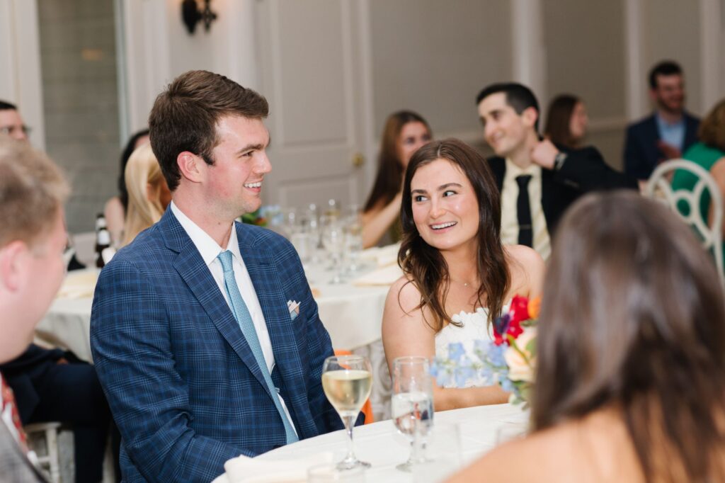 A man in a blue suit and a woman in a white dress sit together at a Chastain Horse Park rehearsal dinner, smiling at each other, while other guests are seated and talking in the background.