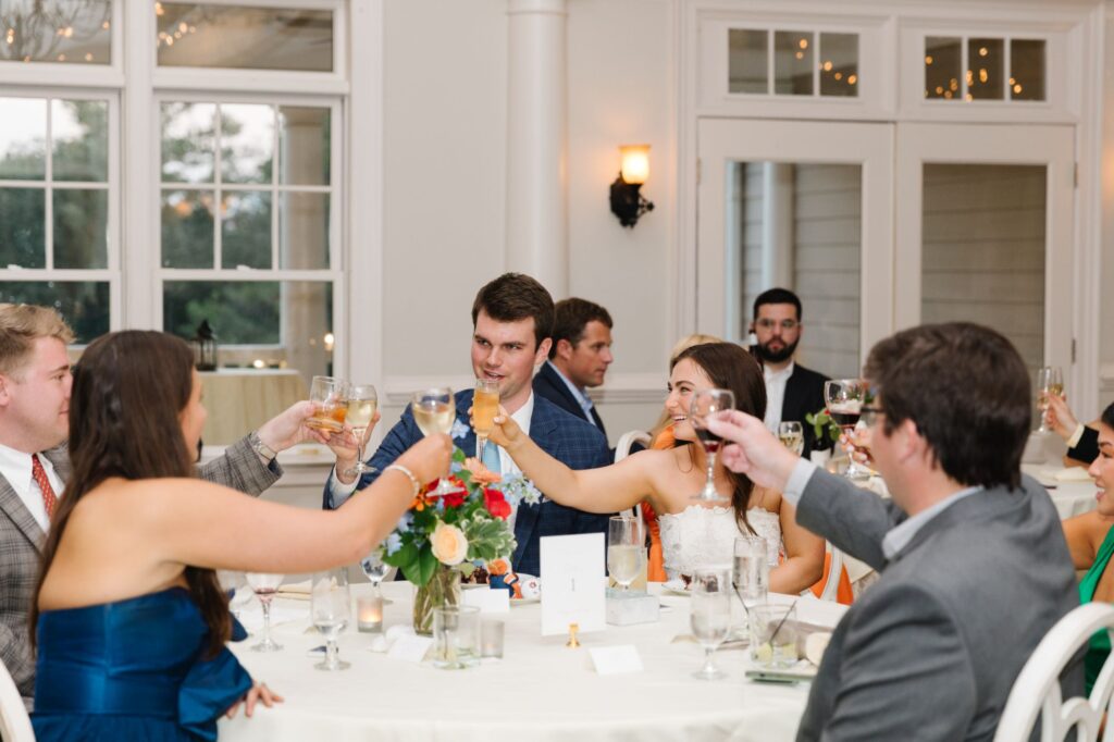 A group of people dressed in formal attire sit around a round table at a Chastain Horse Park rehearsal dinner, raising glasses in a toast. The room is decorated with flowers, candles, and white walls.