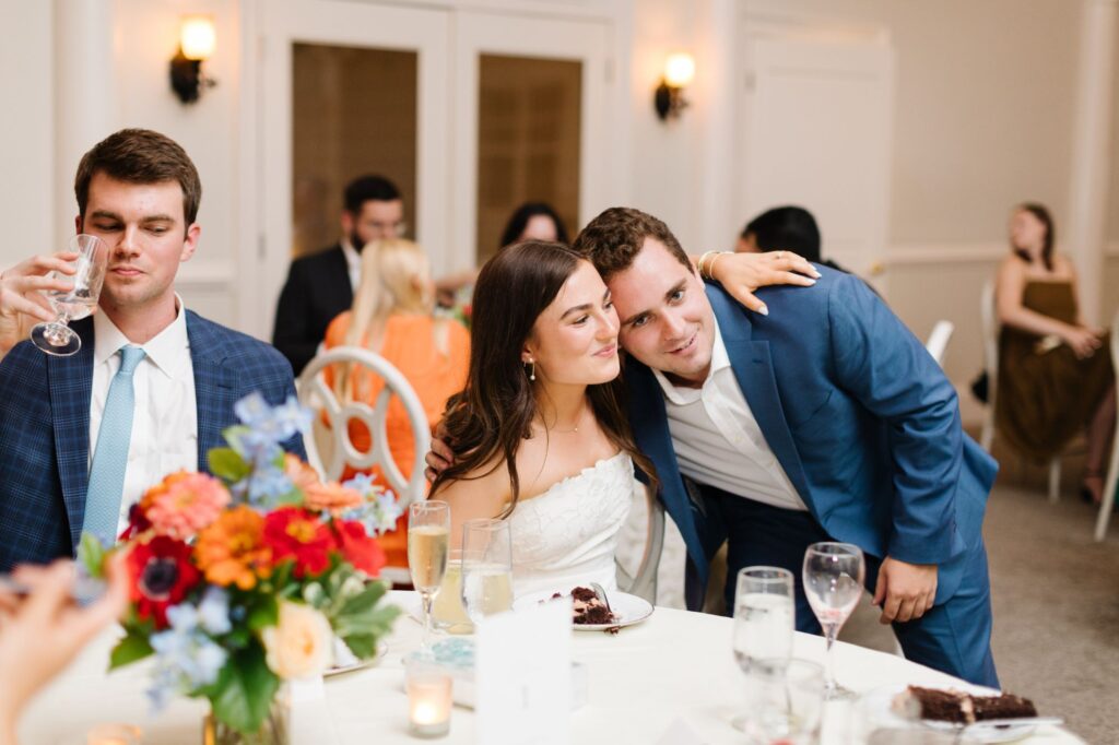 At a Chastain Horse Park rehearsal dinner, a woman in a white dress sits at a table as a man in a blue suit leans in, smiling. Another man enjoys his drink nearby. The table is beautifully set with flowers, drinks, and plates among happy guests.
