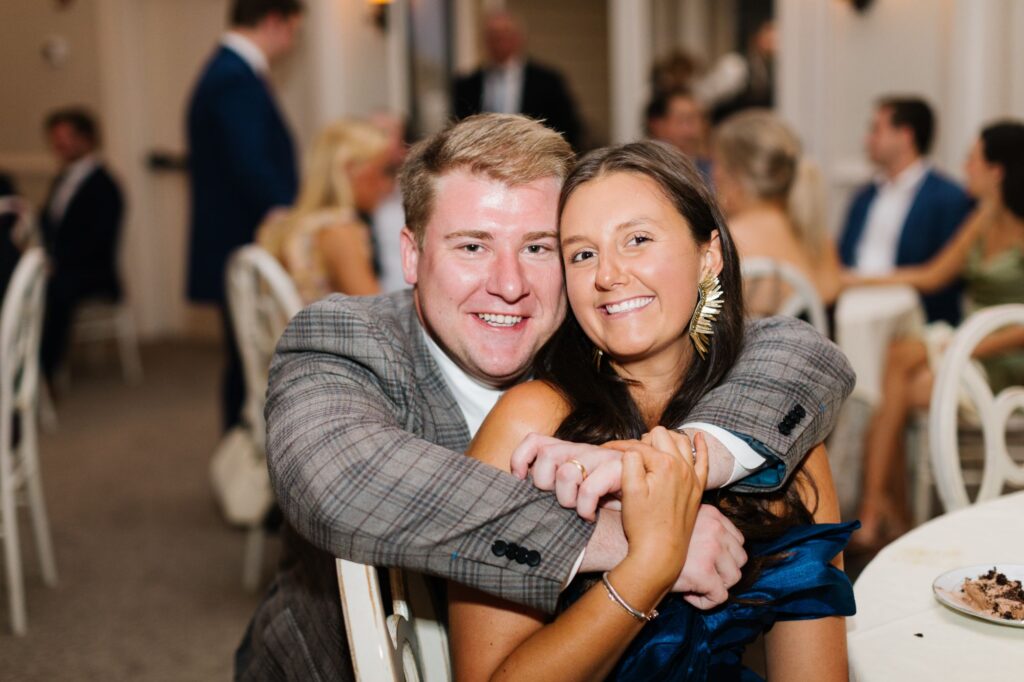 A man in a plaid suit jacket hugs a smiling woman in a blue dress at an indoor Chastain Horse Park rehearsal dinner. They are seated at a table with blurred guests and white chairs in the background.