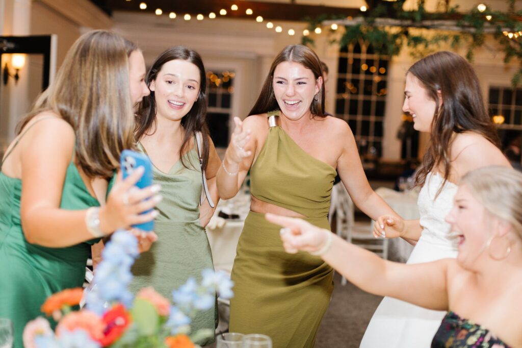 Five women in formal attire smile and laugh together at a brightly lit Chastain Horse Park rehearsal dinner, with one holding a phone for a selfie. Colorful flowers are visible in the foreground.