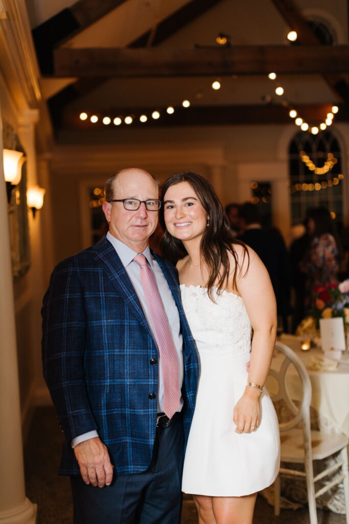 A man in a blue plaid jacket and glasses stands next to a young woman in a white dress, smiling at a Chastain Horse Park rehearsal dinner with string lights and elegant decor in the background.