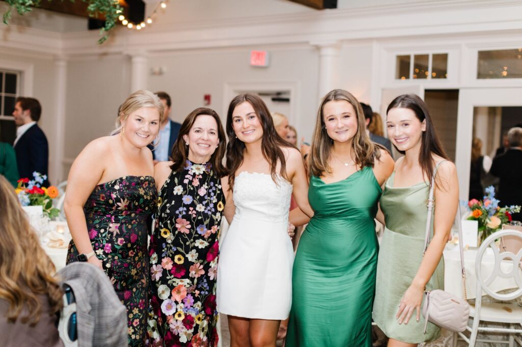 Five women dressed in colorful, semi-formal dresses smile and pose together at a Chastain Horse Park rehearsal dinner, with flowers and other guests visible in the background, creating a joyful celebration atmosphere.