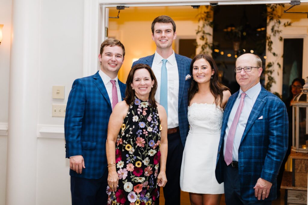 Five people pose indoors at a Chastain Horse Park rehearsal dinner, all dressed formally. Three men wear blue jackets and ties; one woman wears a floral dress, and another wears a white strapless dress. They are smiling and standing close together.