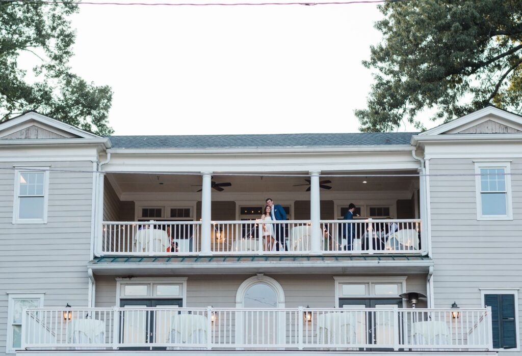 Two people stand on the second-floor balcony of a large, light gray house with white trim at a Chastain Horse Park rehearsal dinner. The house features two levels of balconies, white railings, and is surrounded by tall trees.