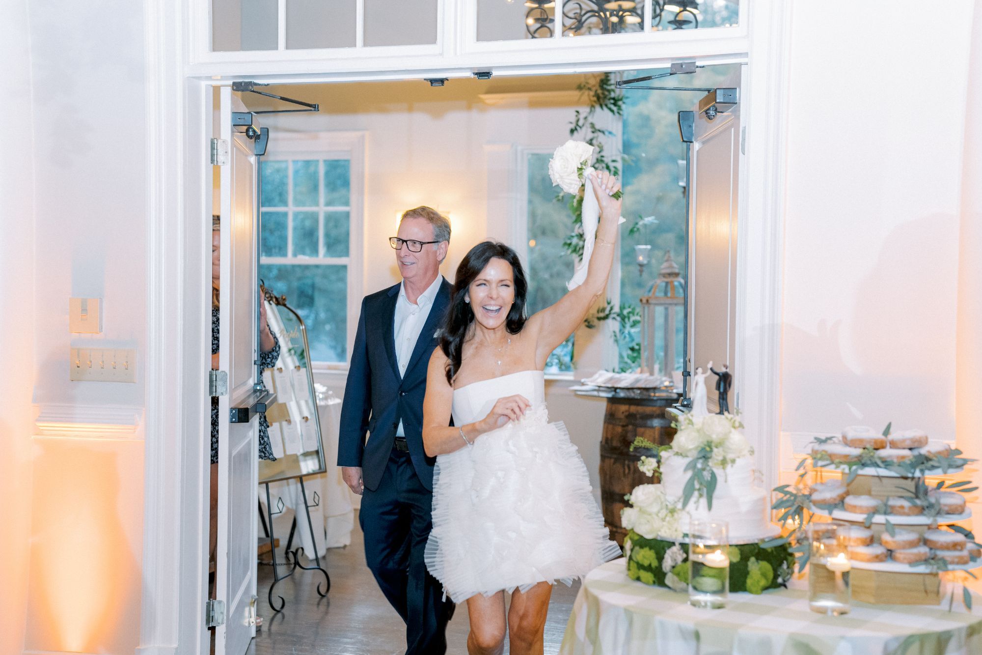 A joyful woman in a short white dress raises a bouquet as she enters a brightly lit room, followed by a smiling man in a suit. The room is decorated with flowers, candles, and a dessert table.