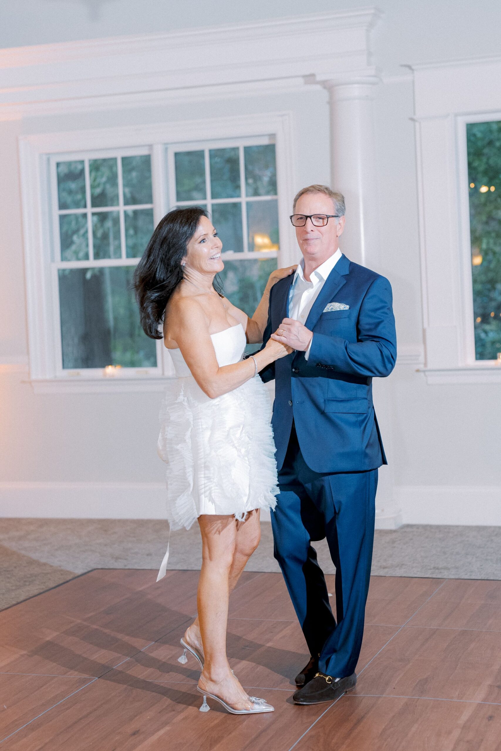 A woman in a short white dress and a man in a blue suit dance together on a wooden floor in a bright room with large windows.