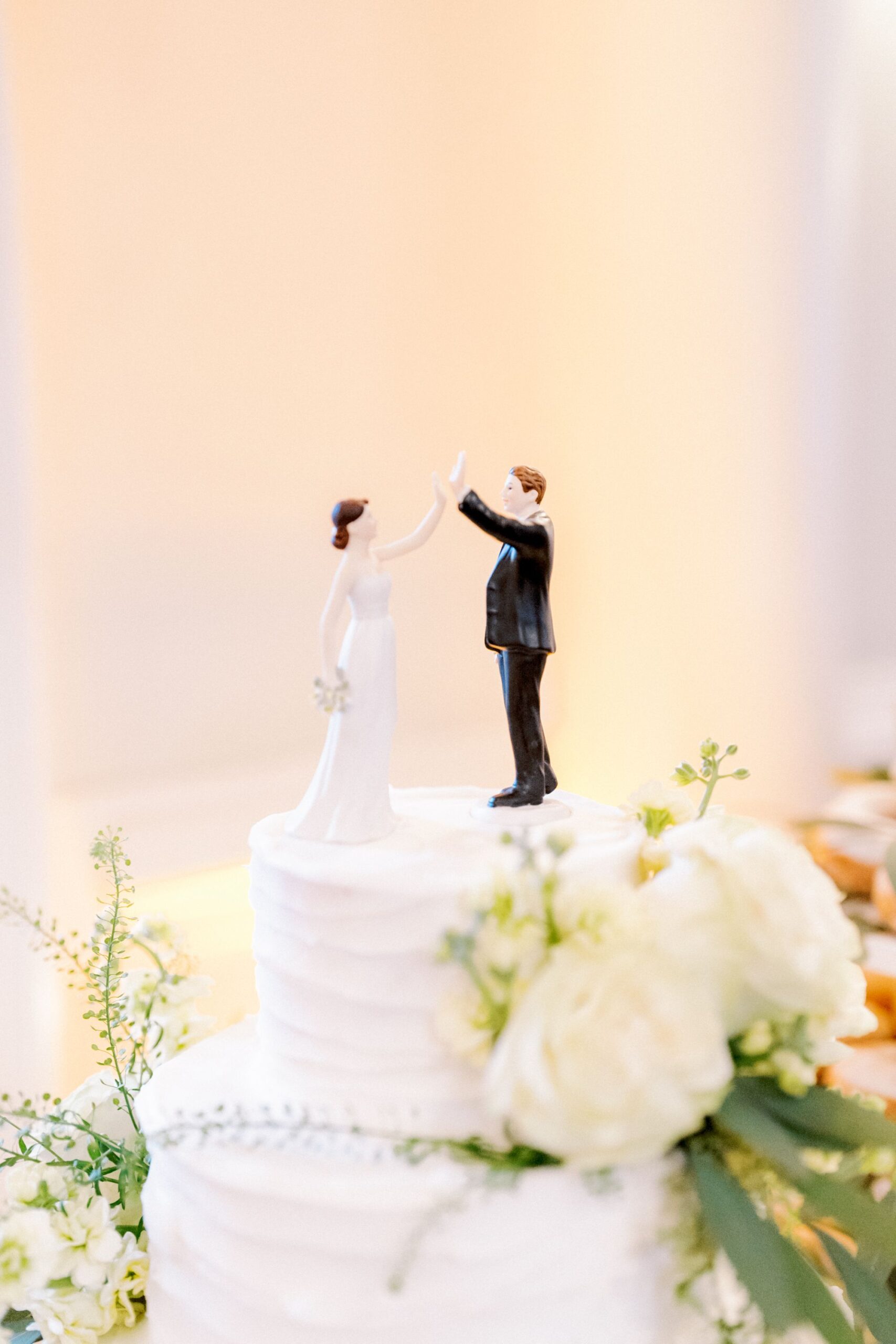 A white wedding cake topped with figurines of a bride and groom giving a high five, decorated with white roses and greenery.