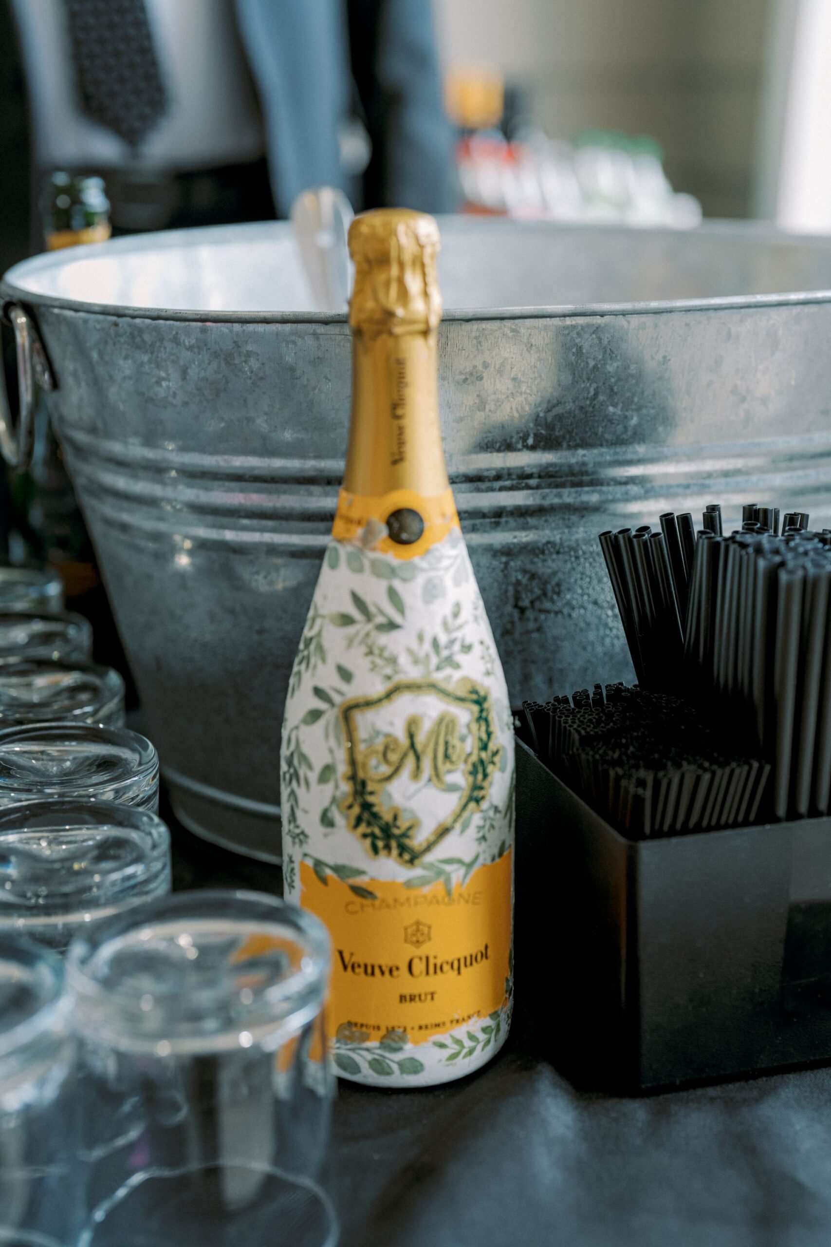 A Veuve Clicquot champagne bottle with a floral design stands on a table next to clear glasses, black straws in a container, and a large metal ice bucket in the background.