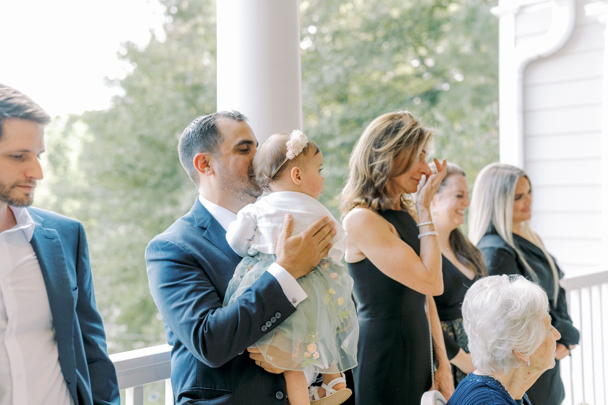 A group of people dressed formally stand on a porch. A man holds a baby in a white dress and headband, while others, including an elderly woman seated in front, smile or wipe away tears. Trees are visible in the background.