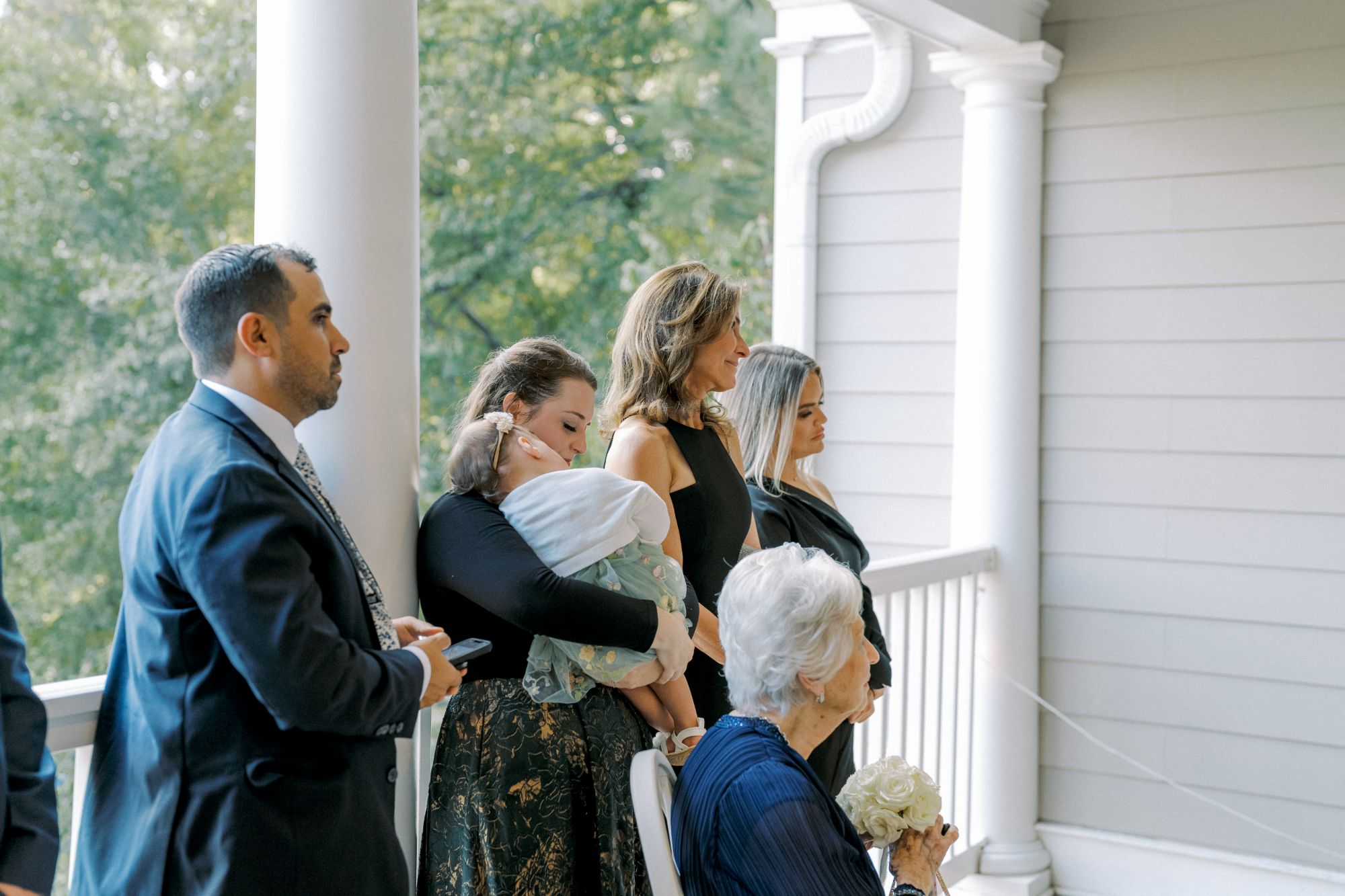 A group of people, including a man in a suit, women in formal dresses, an elderly woman holding flowers, and a woman holding a sleeping baby, stand on a porch with white columns, looking ahead.