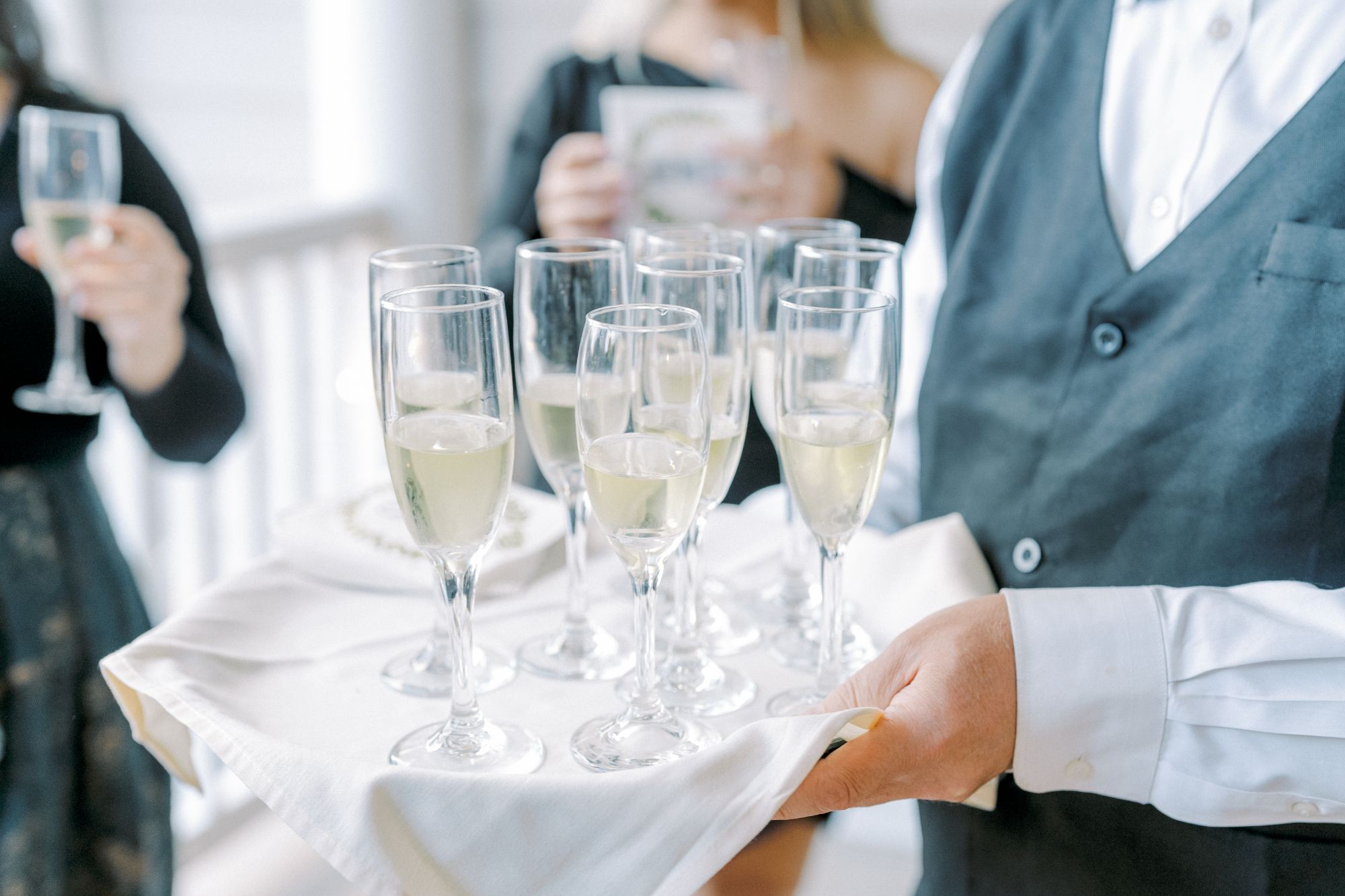 A server in a vest holds a tray with several glasses of champagne. In the background, people are holding drinks and chatting, suggesting a celebratory or formal event.