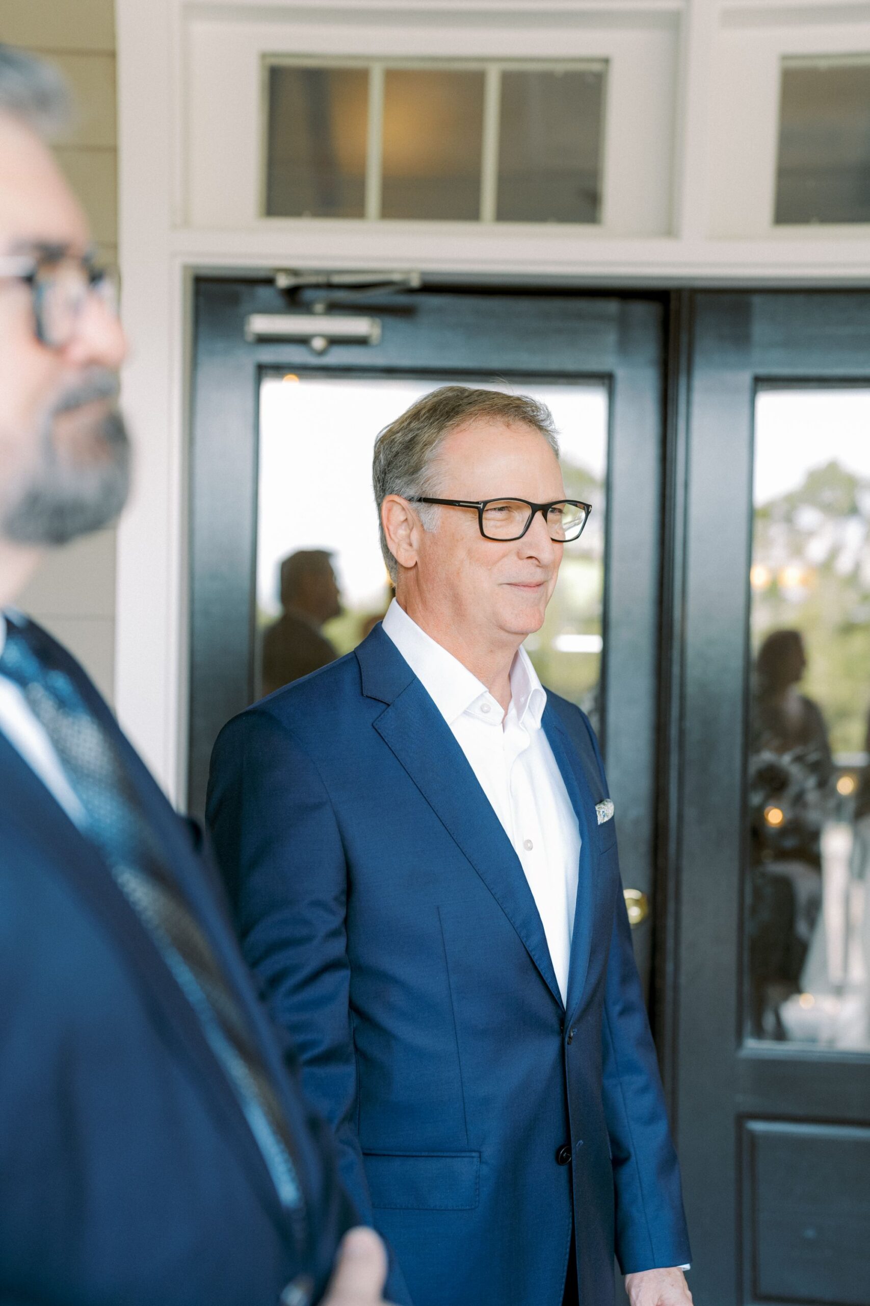 A man wearing a blue suit and glasses stands in front of glass doors, looking ahead. Another person in a suit is partially visible in the foreground, out of focus.
