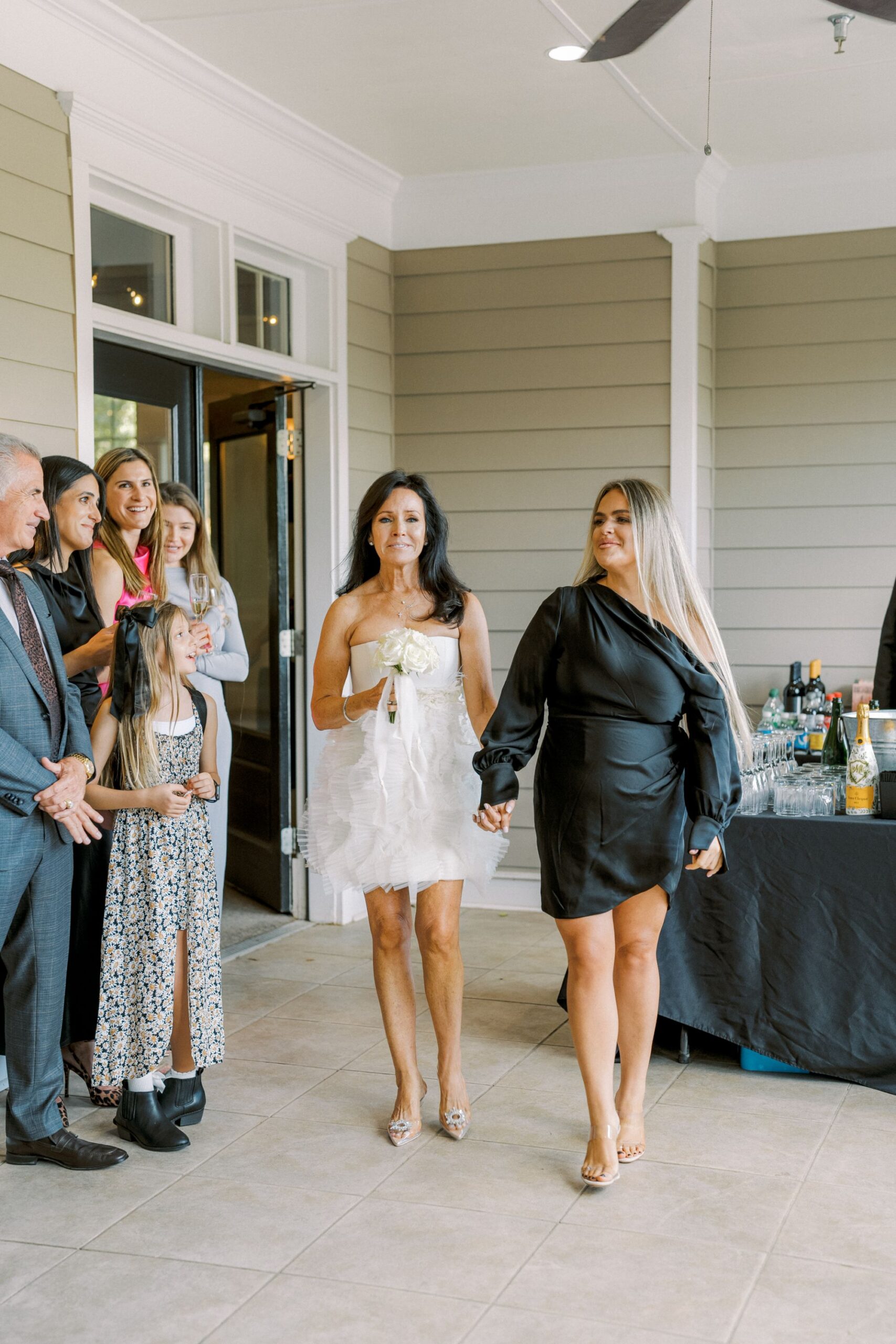 Two women, one in a white ruffled dress and the other in a black dress, hold hands and walk inside a bright room. Several smiling people, including children, stand nearby, and a table with drinks is in the background.
