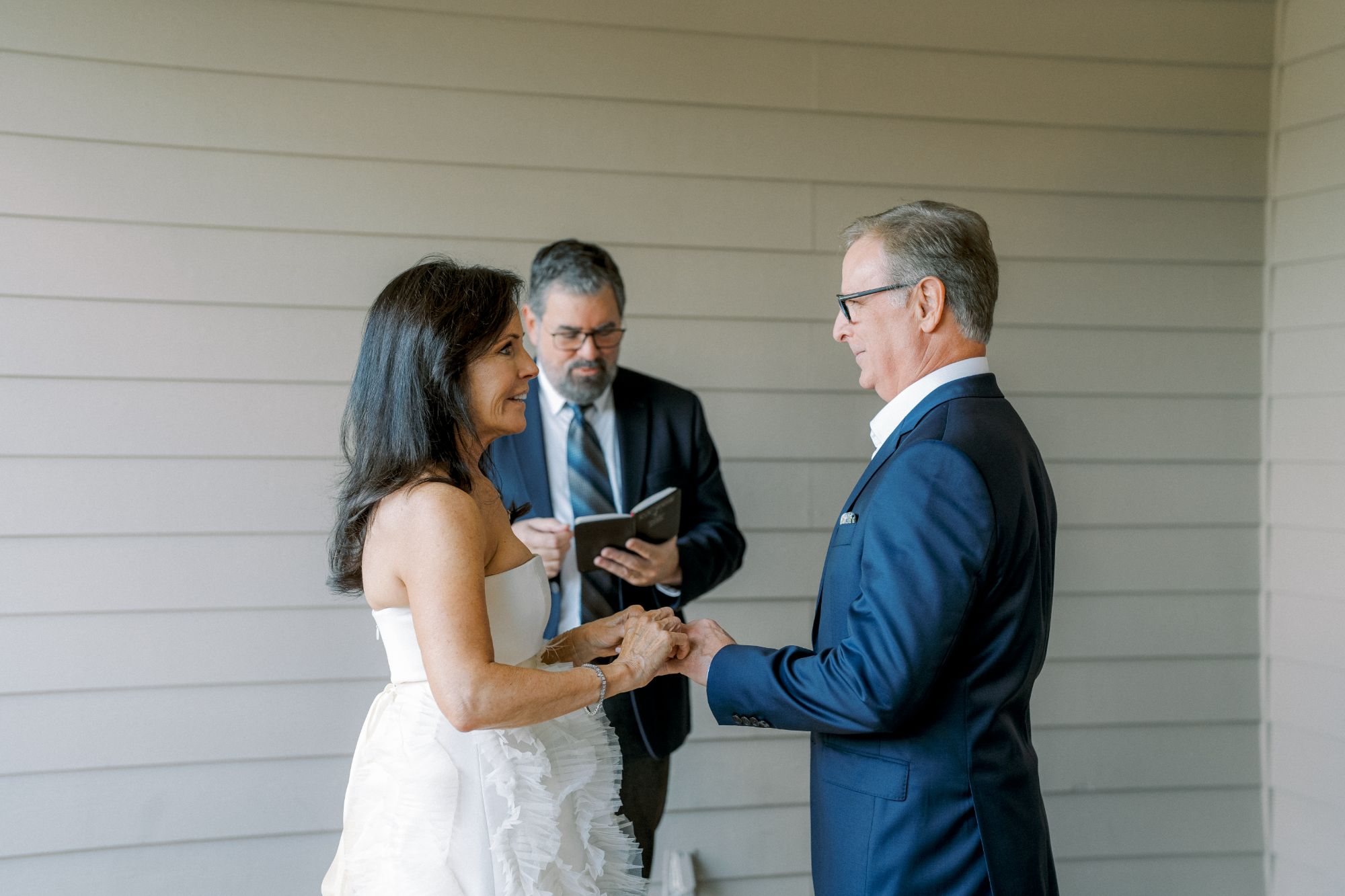 A couple stands facing each other, holding hands, and smiling during a wedding ceremony. An officiant in a suit stands behind them, reading from a book. The setting is indoors with light-colored paneled walls.