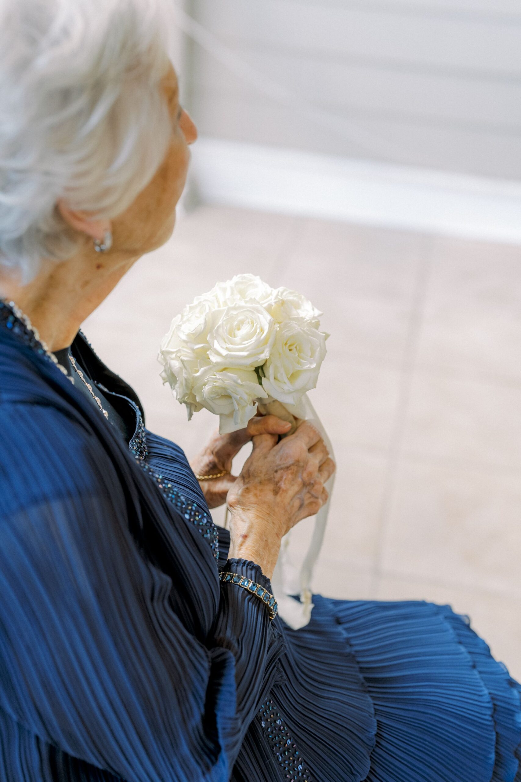 An elderly woman with white hair, wearing a pleated blue dress, sits holding a small bouquet of white roses tied with a ribbon. The background is softly lit and out of focus.