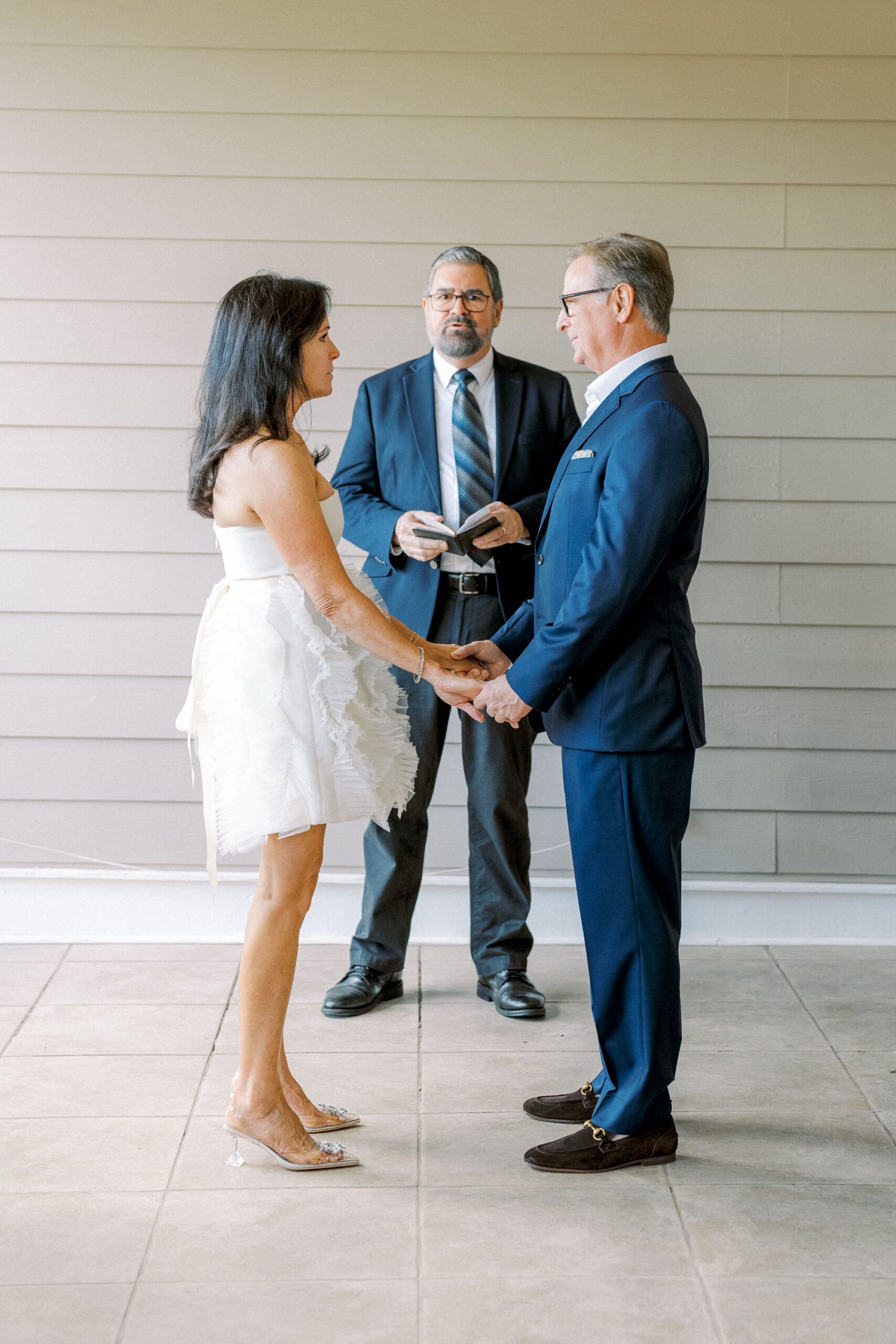 A woman in a white dress and a man in a blue suit stand facing each other, holding hands. A man in a suit stands behind them with a book, appearing to officiate a wedding ceremony indoors.