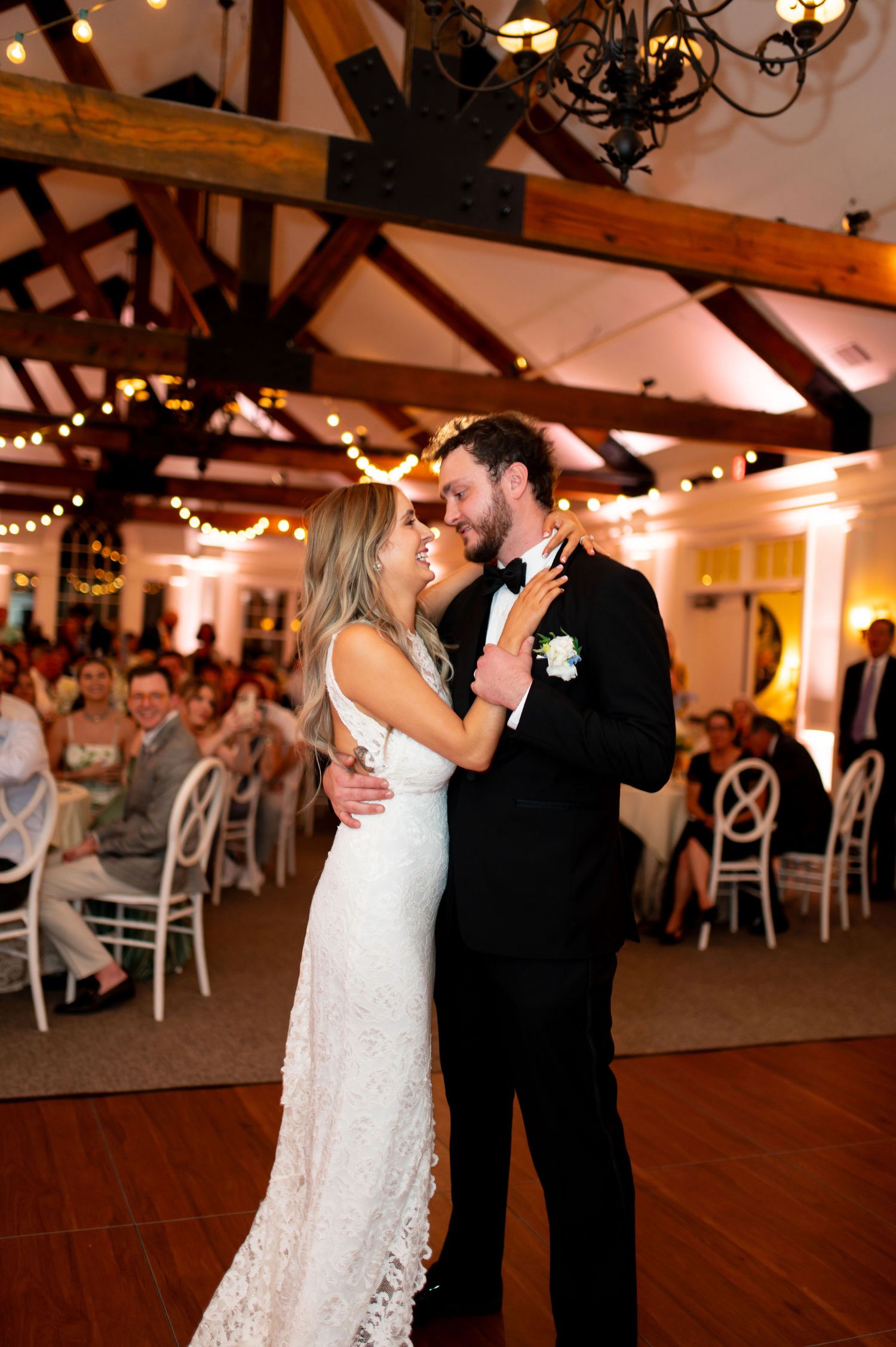 A bride and groom smile at each other while dancing at their wedding reception, surrounded by guests seated at decorated tables under warm string lights and exposed wooden beams during their progressive buffet wedding in Atlanta.