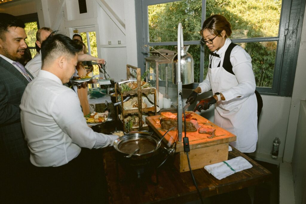 A server in a white apron slices roast beef at a carving station while several guests in formal attire wait in line with plates. A window with greenery outside is in the background.