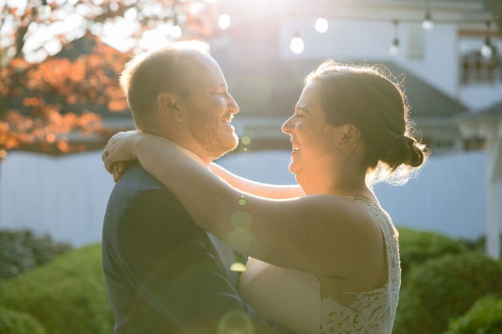 A couple in wedding attire embraces and smiles at each other outdoors, bathed in warm sunlight with a blurred background of trees and a building.