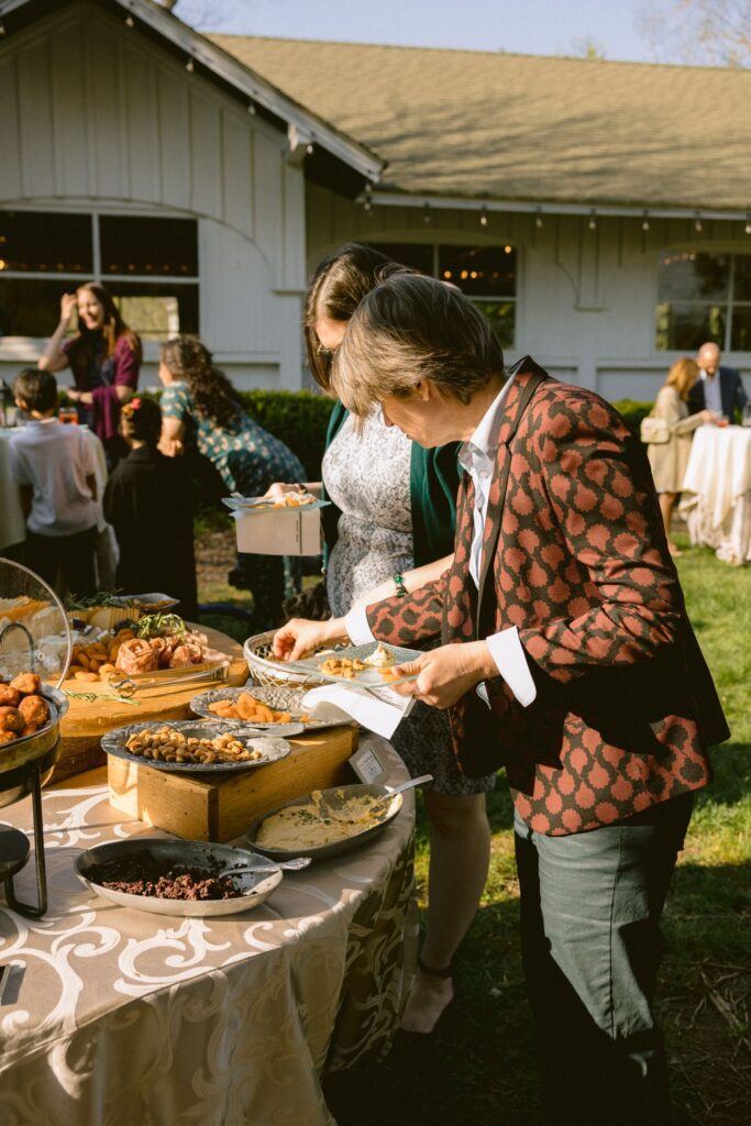 Two people serve themselves food from a buffet table at an outdoor event. Various pastries and dishes are arranged on the table, with other guests and a building visible in the background.