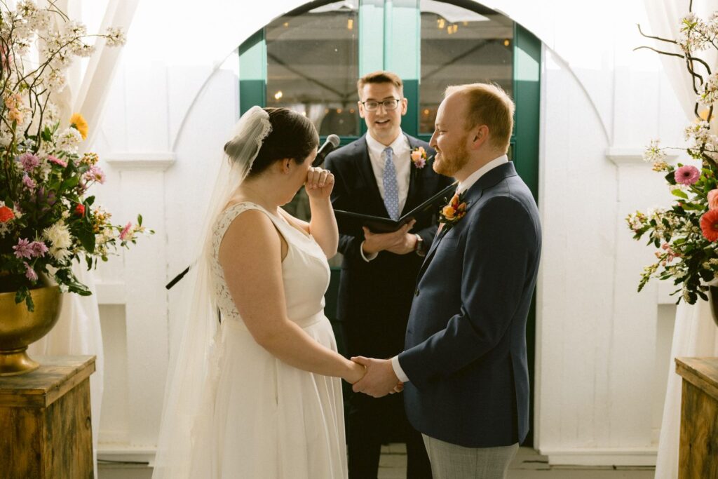 A bride wipes away a tear while holding hands with the groom during their wedding ceremony, with an officiant standing and smiling behind them. Flowers decorate both sides of the scene.