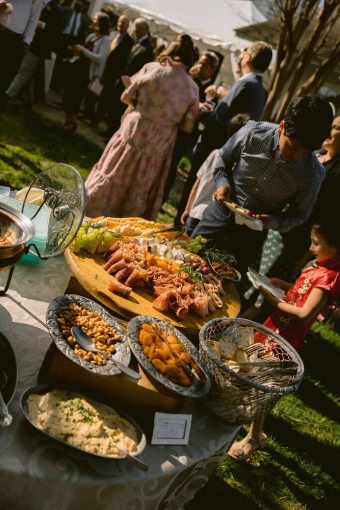 A group of people gather outdoors near a table with assorted appetizers, including meats, cheeses, nuts, and bread, on a sunny day.
