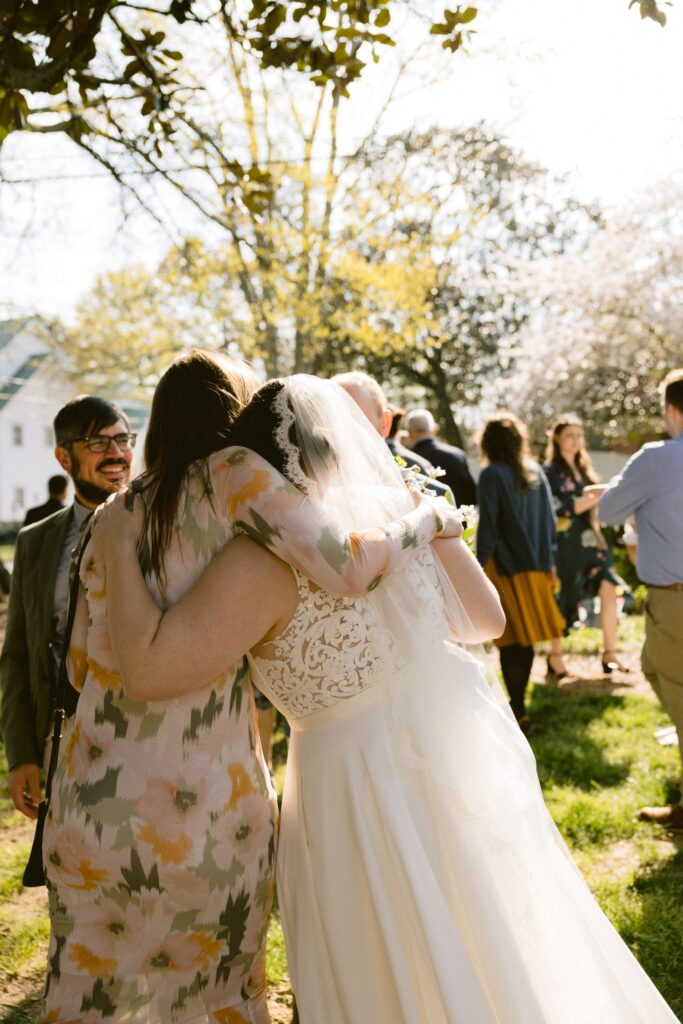 A bride in a white dress and veil hugs a woman in a floral dress outdoors at a sunny wedding, surrounded by guests in a garden setting.