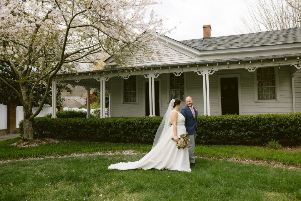A bride in a white gown and veil stands with an older man in a suit outside a white house, surrounded by blooming trees and greenery.