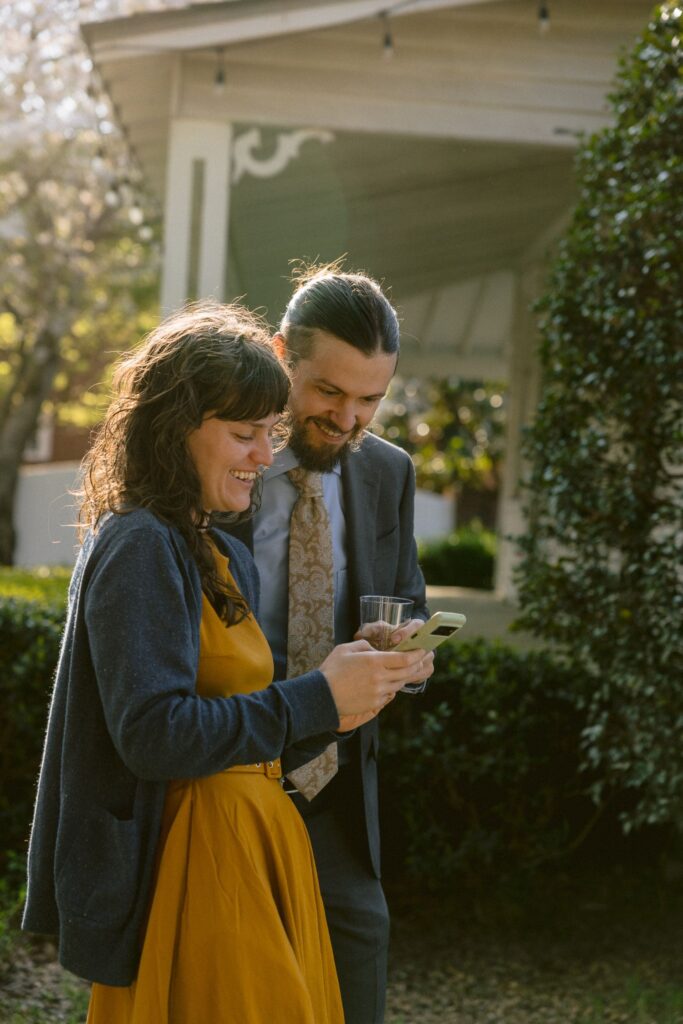 A woman in a yellow dress and a man in a suit stand outside, smiling and looking at a phone together. The man holds a drink. Sunlight filters through the trees near a house.