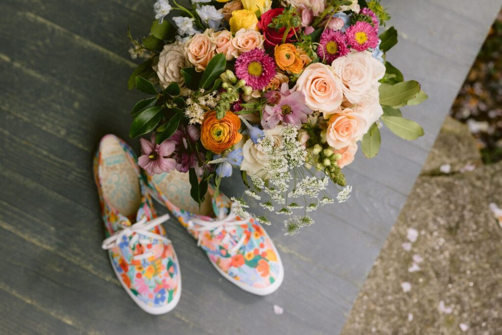 A colorful bouquet of flowers sits on a wooden surface next to a pair of floral-patterned sneakers.