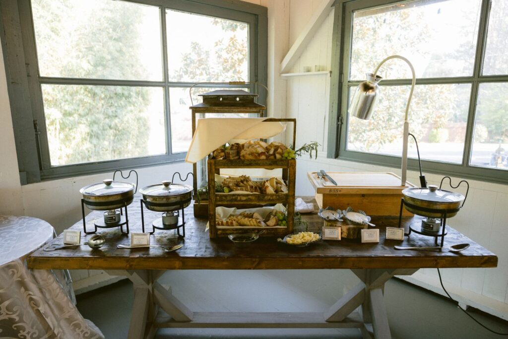 A wooden table with chafing dishes, trays of bread, cheese, and fruit, and utensils set up for a buffet in a bright room with large windows.