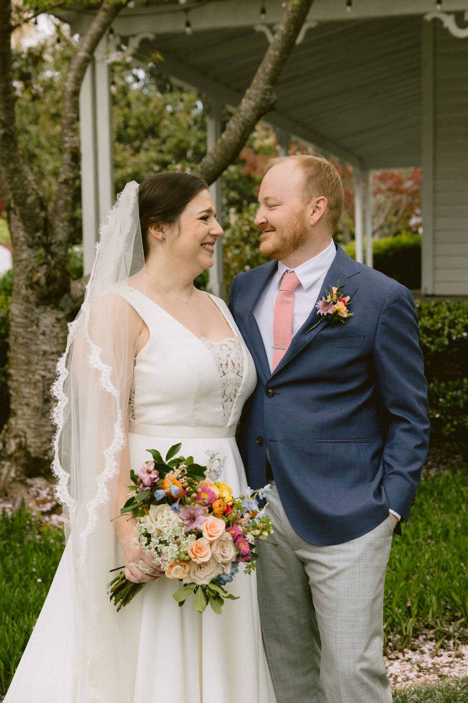 A bride in a white dress holding a colorful bouquet stands smiling beside a groom in a blue suit and pink tie. They look at each other, standing in front of greenery and a white building.