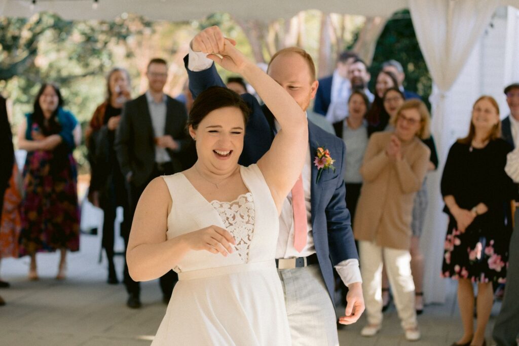 A bride and groom dance together, smiling and surrounded by guests who watch and cheer them on at an outdoor wedding reception.