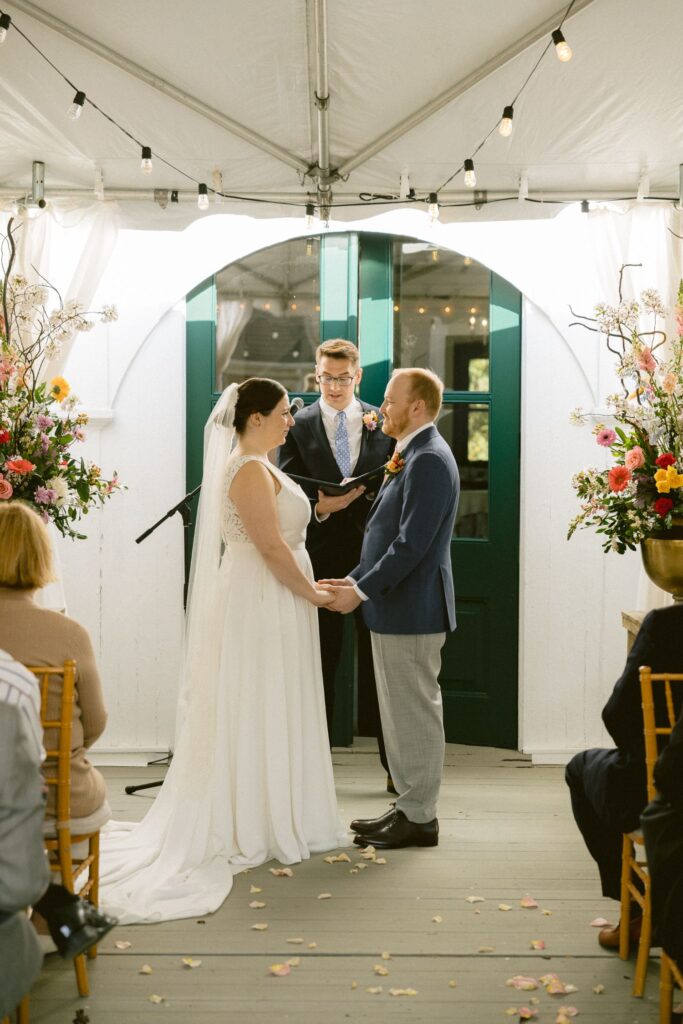 A bride and groom stand holding hands at an indoor wedding ceremony under a tent, with an officiant reading between them. Guests are seated, and colorful flowers decorate the scene.