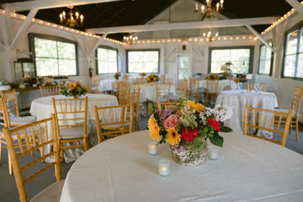A decorated event space with round tables covered in white cloths, wooden chairs, and floral centerpieces. String lights and chandeliers hang from the ceiling, creating a warm, inviting atmosphere.
