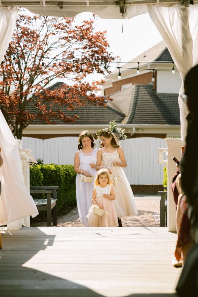 Three young girls in light dresses, holding flowers, walk down an outdoor aisle at a wedding ceremony. Sunlight streams in, and trees and a house are visible in the background.