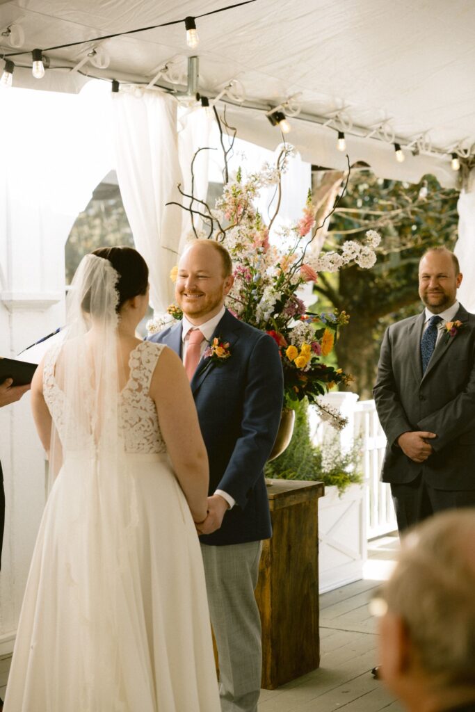A bride and groom stand facing each other, holding hands and smiling, during an outdoor wedding ceremony under a tent, with a groomsman and floral arrangement in the background.
